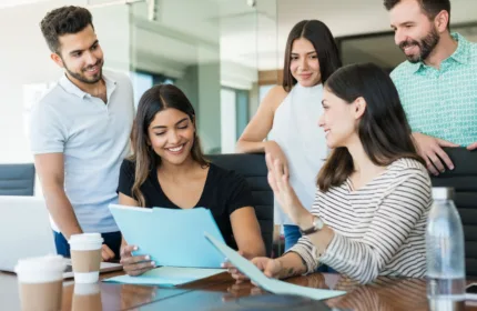 Five professionals gathered around a table reviewing documents and a laptop in an office, representing strategic resourcing and collaboration to maintain operational continuity during companywide restructuring.
