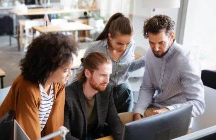 Four professionals gathered around a computer in an office reviewing documents together, representing strategic intervention to optimize accounts payable operations and improve team efficiency.
