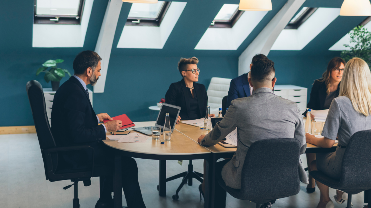 A modern conference room with a round wooden table where several professionals are seated, engaged in discussion. The setting features slanted ceiling beams, large skylight windows allowing natural light, and pendant lights hanging above the table