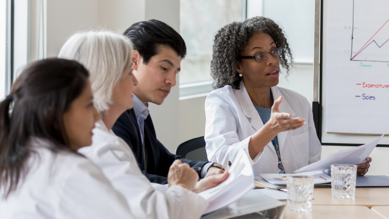 A group of medical professionals seated around a conference table in a bright meeting room, reviewing documents and discussing policies. A flip chart with a line graph is visible in the background, indicating a presentation related to healthcare regulations and compliance.