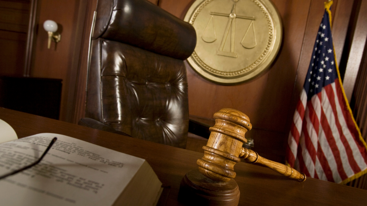 A courtroom scene features a legal professional presenting documents to a judge behind a wooden bench, with law books and traditional furniture, symbolizing litigation.