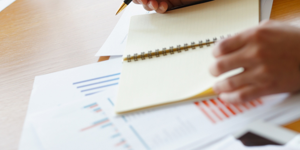 Close-up of a person writing in a notebook while reviewing financial charts and documents related to treasury and cash management on a wooden desk.