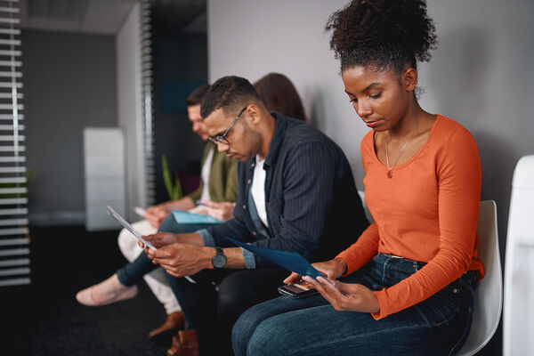 Job candidates sit in a row in an office waiting area, reviewing their resumes before interviews, illustrating common job seeker weaknesses and strategies to improve preparation and confidence.