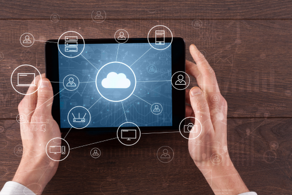 Hands holding a tablet displaying cloud and connected device icons on a wooden surface.